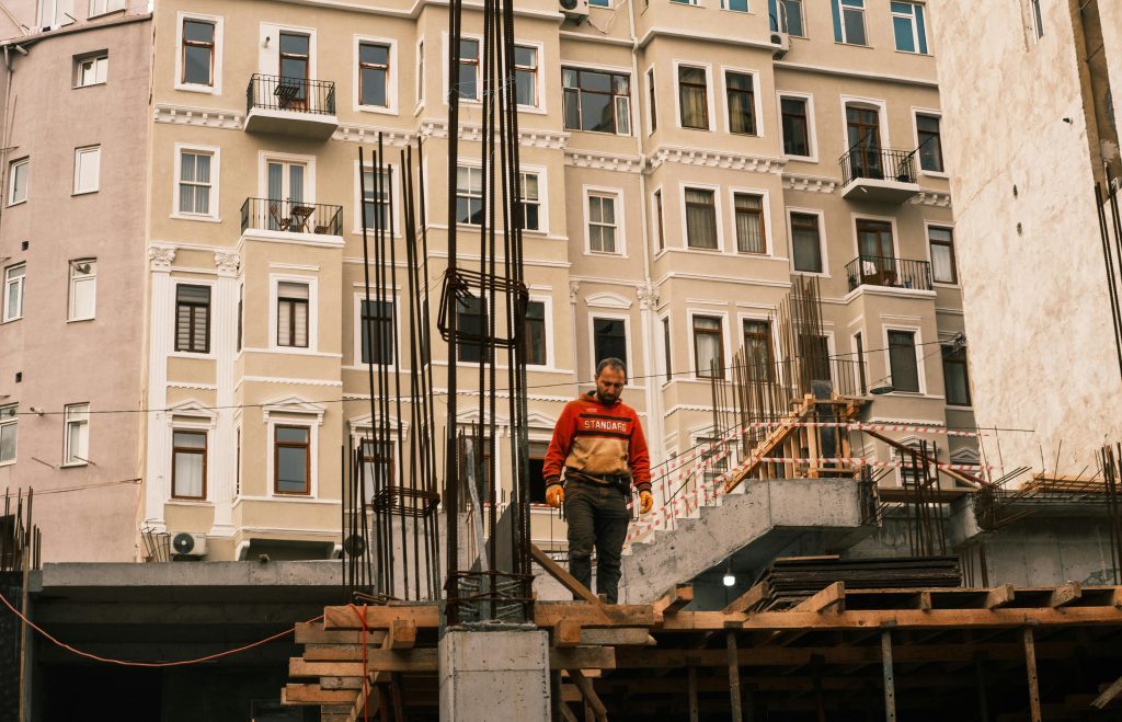 A construction worker stands on a building site in front of a multi-story building facade in a city environment.